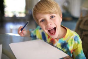 A super happy and excited little boy with a big smile is looking at the camera as he draws with pen on a white paper.